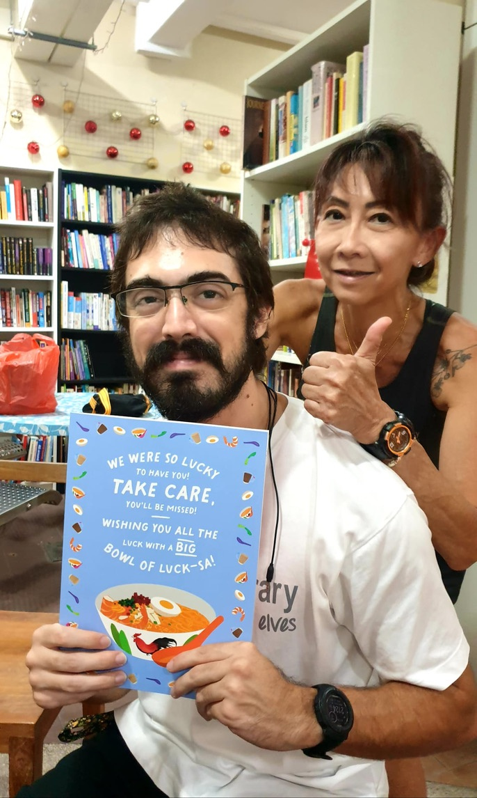 Pedro holding a farewell card beside Cassandra in front of the library shelves