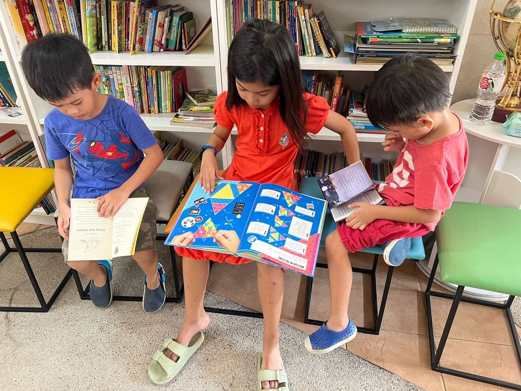 Three children reading books together in front of the library shelves